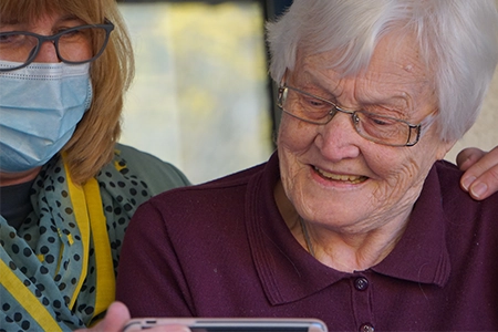 An elderly woman looking at a smart phone. A nurse with a mask is helping her.